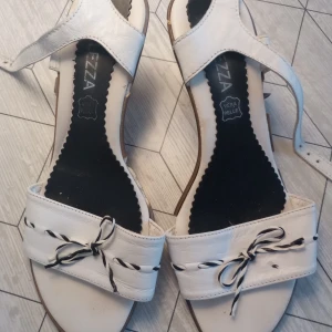 Vita klackskor från Bellezza - Stilrena vita klackskor från Bellezza i läder med dekorativa knytband framtill. Sandalerna har en bekväm passform med justerbara remmar. 