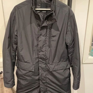 Svart jacka från J.Lindeberg - Snygg svart jacka från J.Lindeberg med dragkedja och knappar. Perfekt för höst och vinter. Jackan har en stilren design med långärmade ärmar och praktiska fickor.