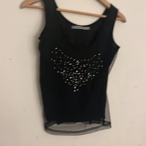 Svart ärmlös topp från Part Two - Snygg svart ärmlös topp från Part Two med dekorativa pärlor och paljetter framtill. Toppen har en transparent nederdel som ger en elegant touch. Perfekt för en kväll ute eller en speciell tillställning.