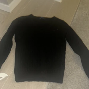 Svart stickad tröja från GANT - Snygg svart stickad tröja från GANT i lammull. Tröjan har en klassisk design med rund hals och ribbade detaljer. Perfekt för kyligare dagar.