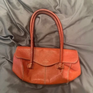 Röd handväska i skinn - Snygg röd handväska i skinn med två långa handtag. Väskan har en dragkedja på insidan och är perfekt för att bära dina dagliga nödvändigheter. Den har en stilren design med synliga sömmar som ger en extra touch. Nån söm som är lös med annars fin.