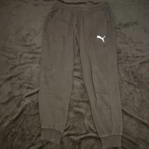 Svarta mjukisbyxor från Puma - Säljer ett par svarta mjukisbyxor från Puma med elastisk midja och ribbade muddar vid bensluten. Byxorna har en vit Puma-logga på ena benet och sidofickor för extra bekvämlighet.