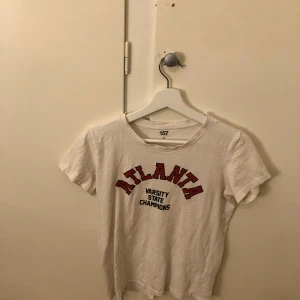 Vit t-shirt med tryck från 157 - Säljer en vit t-shirt från 157 med trycket 'Atlanta Varsity State Champions' i rött och svart. T-shirten är kortärmad och tillverkad i 100% bomull. Perfekt för en avslappnad stil.