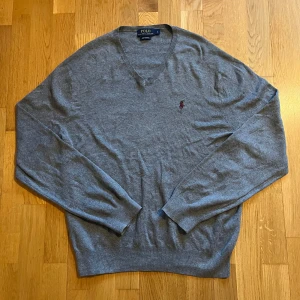 Ralph Lauren tröja - Säljer en stilren grå tröja från Polo Ralph Lauren i fin bomull. Tröjan har en v-ringad design och ett broderat logotypmärke i rött på bröstet. Skick:9/10. Mvh Carl