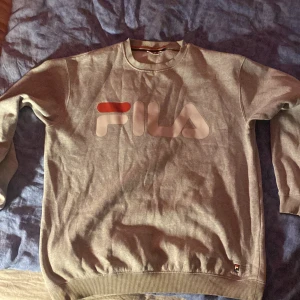 Grå sweatshirt från Fila - Säljer en grå sweatshirt från Fila med stort logotryck på bröstet. Tröjan har långa ärmar och ribbade muddar vid ärmslut och nederkant. Perfekt för en avslappnad stil.