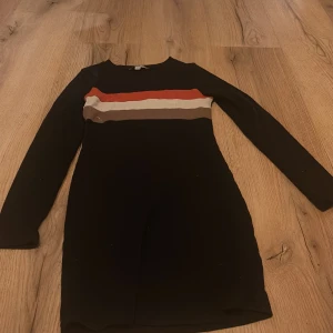 Svart klänning med randigt mönster från Skill - Snygg svart klänning från Skill med långa ärmar och ett coolt randigt mönster i rött, beige och brunt över bröstet. Perfekt för höst och vinter med sitt stilrena och bekväma design. Passar utmärkt för både vardag och speciella tillfällen.