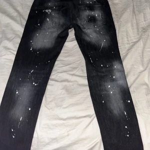 Svarta jeans med slitningar från Dsquared2 - Snygga svarta jeans från Dsquared2 med coola slitningar och färgstänk. De har en modern, tight passform och är perfekta för en trendig look. Jeansen har klassisk femficksdesign och knappgylf. AA kopior. Säljer för att jag blev lurad själv. Köpte de för 2500. Knappt använda. 