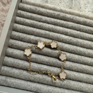 Guldarmband med blommotiv - Säljer ett elegant guldarmband med fem vita blommor. Armbandet har en fin kedja och ett säkert lås. Perfekt för att ge en touch av lyx till vilken outfit som helst. Passar både till vardag och fest!