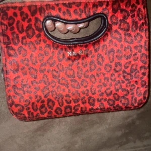 Röd leopardmönstrad väska från NA-KD - Säljer en supercool röd leopardmönstrad väska från NA-KD. Den har ett praktiskt handtag. Väskan är i syntetmaterial och har en stilren design som passar både till vardags och fest. 