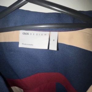 Randig kortärmad skjorta från ASOS DESIGN - Säljer en snygg kortärmad skjorta från ASOS DESIGN i storlek M. Skjortan har ett coolt randigt mönster i blått, beige och rött. Perfekt för sommaren eller en avslappnad fest. Den har knappar framtill och är gjord i ett lätt och bekvämt material. Passar perfekt för en stilren och avslappnad look!