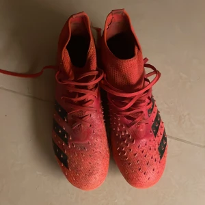 Röda Adidas Predator fotbollsskor - Säljer ett par coola röda Adidas Predator fotbollsskor i bra skick. De har en snygg design med svarta detaljer och snörning. Perfekta för fotbollsspelare som vill ha bra grepp och stil på planen. Skorna har en tight passform och är redo för nästa match!