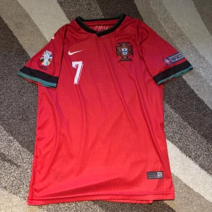 Röd Portugal fotbollströja - Säljer en röd Portugal fotbollströja från Nike med nummer 7 och namn på baksidan. Tröjan har korta ärmar och är i Dri-FIT material för bra komfort. Perfekt för fotbollsfans! 🇵🇹