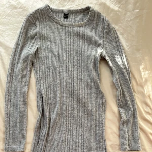 Grå ribbad långärmad tröja - Säljer en stilren grå ribbad tröja med långa ärmar. Perfekt för höst och vinter, den är både bekväm och snygg. Tröjan har en unik design med en slits framtill. 