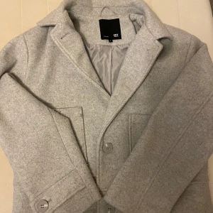 Grå kappa från 157 - Säljer en stilren och klassisk grå kappa från 157. Den har en normal passform med långa ärmar och knappar framtill. Perfekt för höst och vår när du vill hålla dig varm och snygg. 🧥