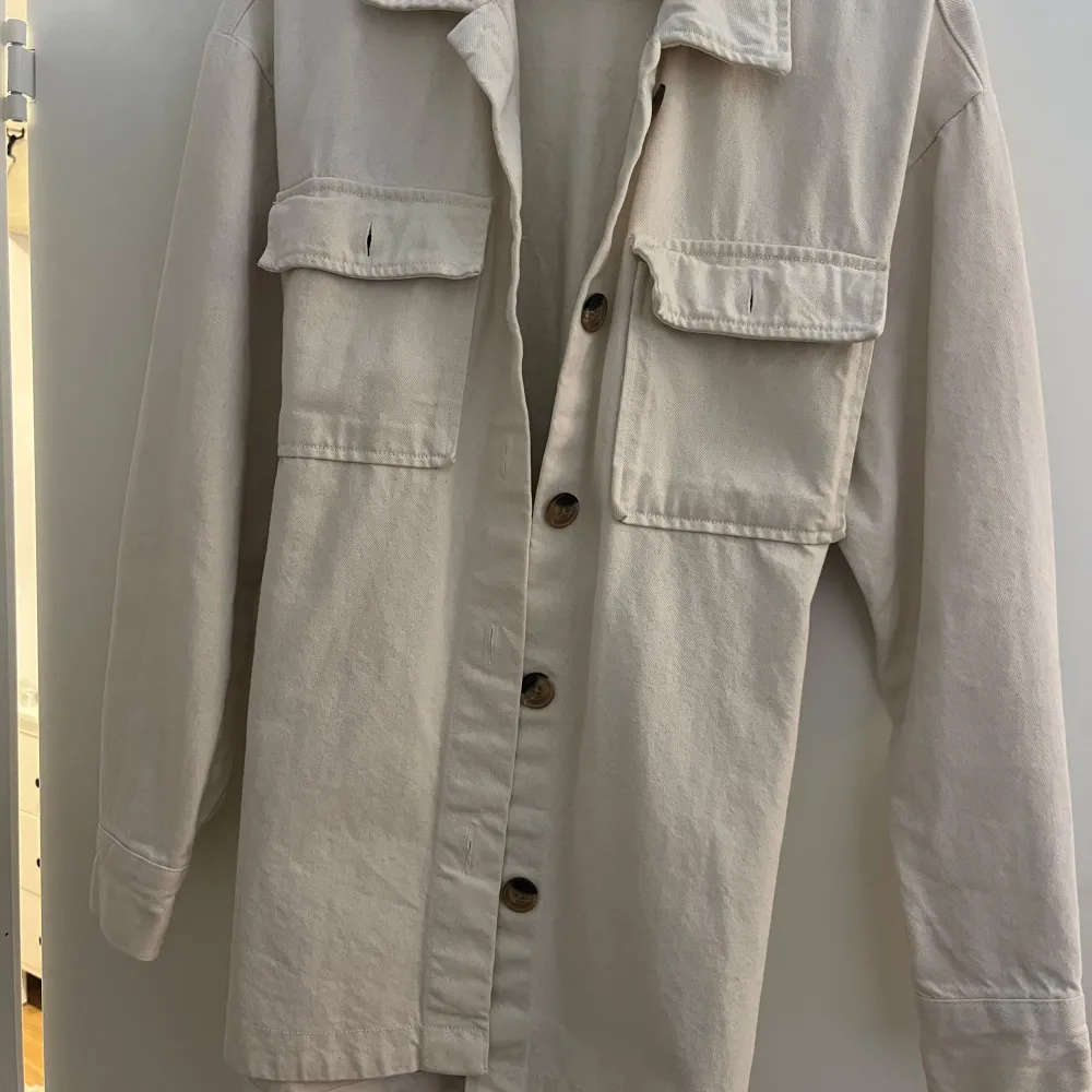 Säljer en superfin beige overshirt från Bik Bok i storlek XS. Den har långa ärmar och knappar framtill, samt två stora fickor på bröstet. Materialet känns som bomull och den har ett bälte i midjan för en snygg passform. Perfekt nu till höst!. Takit.