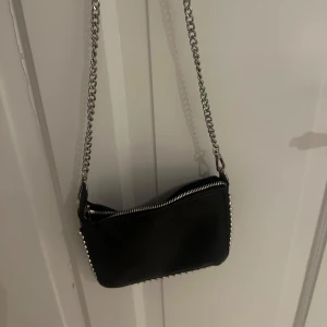 Svart axelväska med kedja - Säljer en snygg svart axelväska med en cool kedja som axelrem. Väskan har en dragkedja upptill och är perfekt för en utekväll eller när du vill ha med det viktigaste. Den är i skinn och har en stilren design med nitar längs kanterna. Passar till alla outfits!
