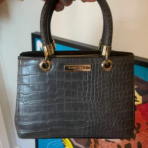 Kurt geiger handväska - Superfin kurt geiger väska med gulddetaljer och krokodil liknande mönster Tillhörande axelband finns Dragkedjan har tyvärr gått sönder(därav priset)men det finns knappar som man kan stänga med så inget som påverkar självaste funktionen😊