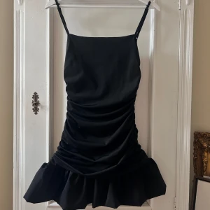 Svart klänning med volangdetalj - Säljer en elegant svart klänning med smala axelband och en snygg volangdetalj längst ner. Klänningen har en åtsittande passform med rynkade detaljer som ger en smickrande siluett. Perfekt för festliga tillfällen!
