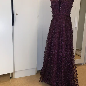 Mörklila långklänning med spetsdetaljer - Säljer en fantastisk mörklila långklänning med v-ringning och vackra spetsdetaljer. Klänningen har en elegant passform och är perfekt för festliga tillfällen. Den är dekorerad med små knappar framtill och har en fin struktur som ger en lyxig känsla.