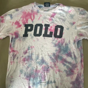 Tie-dye t-shirt från Polo Ralph Lauren - Säljer en supercool tie-dye t-shirt från Polo Ralph Lauren. Den har en vit bas med rosa och blå mönster och ett stort svart 'POLO' tryck på framsidan. Perfekt för en avslappnad stil! Finns inte i affärer!!