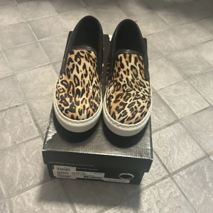 Leopardmönstrade slip-ons från Nilson Shoes - Säljer ett par coola slip-ons från Nilson Shoes med leopardmönster. Skorna har en vit sula och är i nyskick. Perfekta för att ge din outfit en extra edge! 🐆
