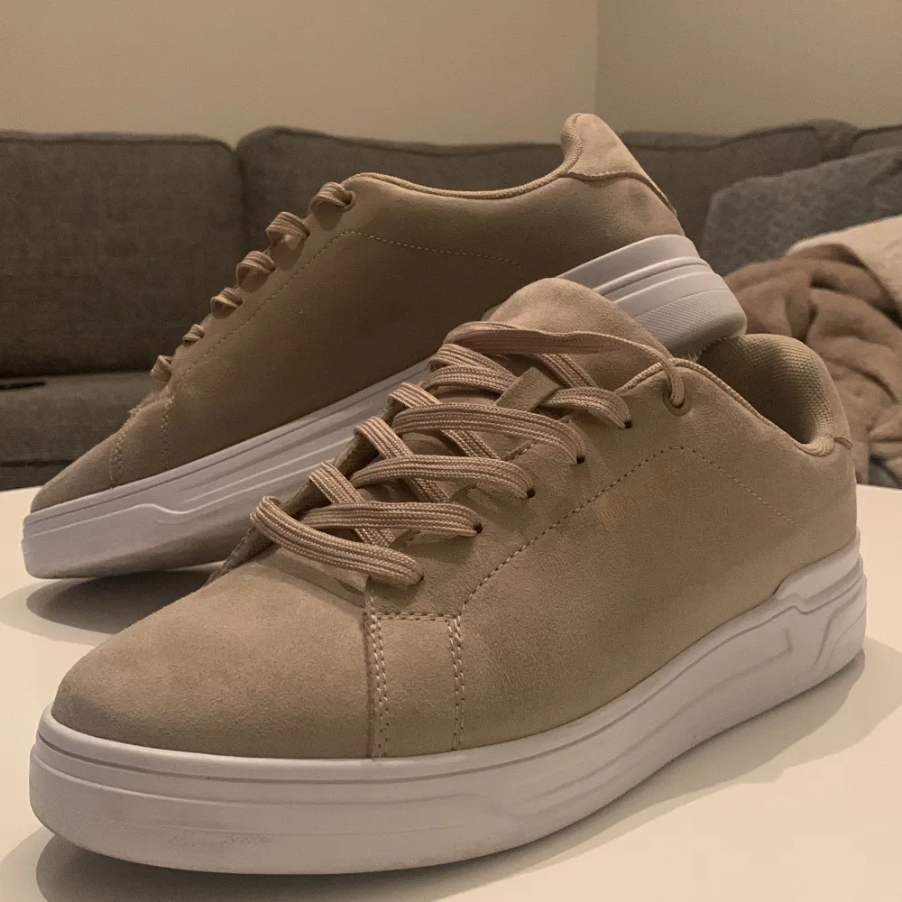 Säljer ett par snygga och stilrena beige sneakers från Iguassu. De har en klassisk design med snörning och en vit sula som ger en fräsch look. Perfekta för vardagsbruk och passar till det mesta i garderoben. Skorna är i storlek 41 och är i nyskick.. Skor.