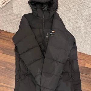 Svart pufferjacka från Sail Racing - Säljer en svart pufferjacka från Sail Racing i storlek L. Jackan är perfekt för kalla dagar med sin tjocka vaddering och huva. Den har en dragkedja framtill och justerbara snören vid ärmsluten för extra skydd mot kyla. Passar perfekt för både vardag och utomhusaktiviteter. Håll dig varm och stilren i vinter!