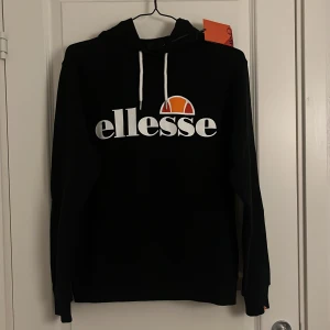 Svart hoodie från Ellesse - Säljer en snygg och bekväm svart hoodie från Ellesse. Den har en stor logga på framsidan i vitt och orange, och en praktisk känguruficka. Perfekt för en avslappnad stil eller för att hålla sig varm under kyliga dagar. Huvan har vita dragsnören för justering. Helt ny med tag kvar!
