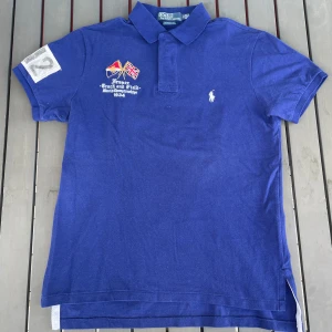 Blå pikétröja från Ralph Lauren - Säljer en snygg blå pikétröja från Ralph Lauren. Den har korta ärmar och en klassisk krage. På framsidan finns broderade flaggor och texten 'France Track and Field March Championships 1934'. Baksidan har texten 'FRANCE' och siffrorna '012'. Perfekt för en sportig look!