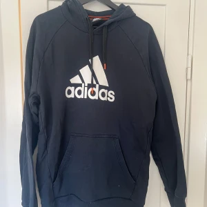 Svart hoodie från Adidas - Säljer en svart hoodie från Adidas med deras klassiska logga i vitt på framsidan. Den har en stor ficka framtill och justerbar huva med snören. Perfekt för en avslappnad stil eller träning. Skön och bekväm att ha på sig!