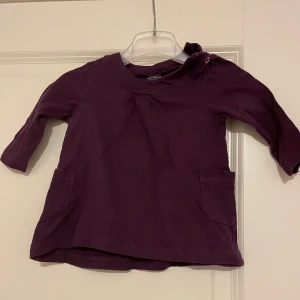 Lila långärmad babyklänning storlek 74 - Söt lila långärmad klänning för baby med två fickor framtill. Oanvänd.