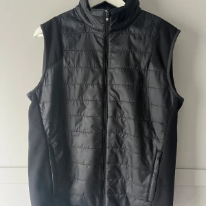 Svart väst från FootJoy - Snygg svart väst från FootJoy med dragkedja framtill. Västen har en quiltad design och är perfekt för kyligare dagar. Den är ärmlös och har en stilren look som passar till många olika outfits.