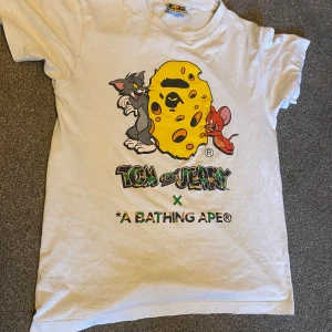Vit Tom and Jerry x A Bathing Ape t-shirt. Bape T-shirt  - Cool vit t-shirt med Tom and Jerry-motiv i samarbete med A Bathing Ape. Designen visar Tom och Jerry med bape inspirerande föremål. Bape t shirten är perfekt för sommaren 🎉⚡️