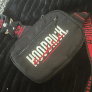 Svart axelväska från Hoodrich - Säljer en svart axelväska från Hoodrich med röd och vit logga. Väskan har en justerbar rem med text och en praktisk dragkedja framtill. Perfekt för att bära med sig det nödvändigaste.