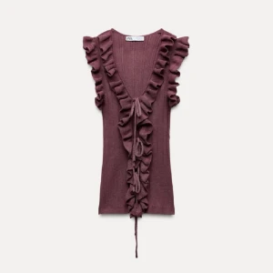 Vinröd topp med volang från Zara - Säljer en vinröd topp från Zara med volangdetaljer längs den v-ringade halsen. Toppen har en ribbad struktur och knytband framtill. Den är helt ny med prislappen kvar. Skriv om frågor