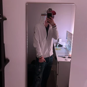 Vit hoodie från Moncler - Säljer en stilren vit hoodie från Moncler med dragkedja och huva. Den har snygga randiga detaljer i rött och svart på ärmsluten och huvan. Perfekt för en casual look med en touch av lyx kom med bud storlek s. Vet ej äktheten då jag köpte den för 1100 på vinted och den ej passade däravpriset