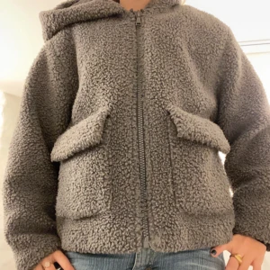 Grå teddyjacka med huva - Mysig grå teddyjacka med huva och dragkedja framtill. Jackan har två stora fickor och är perfekt för kyliga dagar. Den är långärmad och har en avslappnad passform. I en av fickorna finns det ett hål och det är därför jag säljer billigare😊Jackan passar mig som är 160!