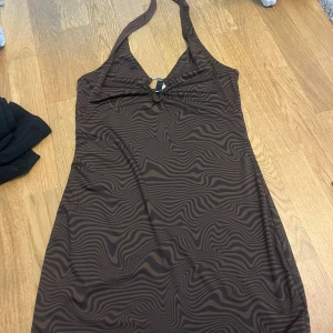 Brun mönstrad klänning med halterneck - Snygg brun klänning med ett unikt vågigt mönster och halterneck-design. Klänningen har en dekorativ ringdetalj framtill som ger en extra touch. Perfekt för en stilfull look.