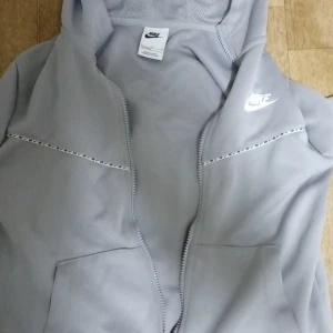 Nike tech - Säljer en stilren grå hoodie från Nike med dragkedja och huva. Den har ett diskret Nike-logotyptryck på bröstet och dekorativa sömmar. Perfekt för en avslappnad look.