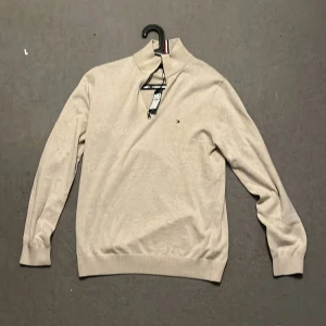 Beige tröja från Tommy Hilfiger - Säljer en stilren beige tröja från Tommy Hilfiger med dragkedja och liten logga på bröstet. Tröjan har långa ärmar och ribbade muddar. Perfekt för en avslappnad och snygg look. Helt oanvänd!!! Farsen köpte den i USA men den passa inte riktigt och vi kan ej lämna tillbaka den därför jag säljer. Ordnarie pris runt tusen lapen