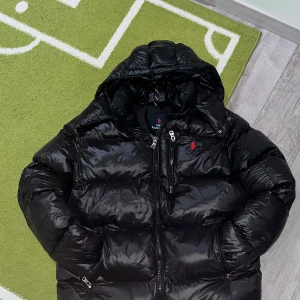 Svart dunjacka från Ralph Lauren - Snygg svart dunjacka från Ralph Lauren med huva och dragkedja. Perfekt för kyliga dagar. Jackan har en klassisk design med diskret logga på bröstet. Helt ny och oanvänd