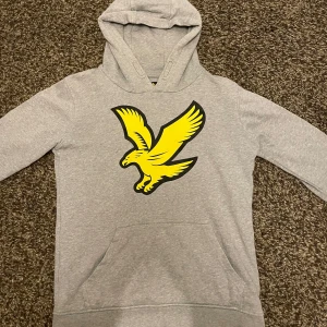 Grå hoodie från Lyle & Scott - Säljer en grå hoodie från Lyle & Scott med en stor gul lyle & scott märke på framsidan. Tröjan har en klassisk känguruficka. Perfekt för en avslappnad stil. 