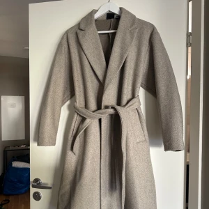 Beige kappa från Vero Moda - Snygg beige kappa från Vero Moda med klassisk design och knytskärp i midjan. Perfekt för kyligare dagar med sin stilrena look och långa ärmar. Ett måste i garderoben för den som älskar tidlös stil.