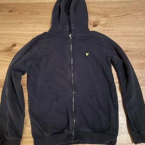 Svart huvtröja från Lyle & Scott - Säljer en mörkblå huvtröja från Lyle & Scott med dragkedja och en broderad logotyp på bröstet. Tröjan har långa ärmar och en praktisk huva. Perfekt för en avslappnad stil.