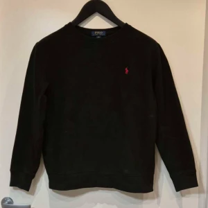Svart tröja från Polo Ralph Lauren - Säljer en stilren svart tröja från Polo Ralph Lauren med deras ikoniska röda logga på bröstet. Tröjan har långa ärmar och en klassisk rund halsringning. Perfekt för en avslappnad och snygg look. Storlek är 14-16 och L.