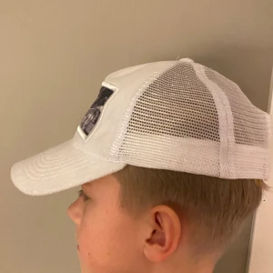 Vit keps med fotomotiv - Snygg vit keps från Berretta med ett coolt fotomotiv på framsidan. Kepsen har ett nätmaterial på sidorna och justerbar passform. Perfekt för en avslappnad stil.