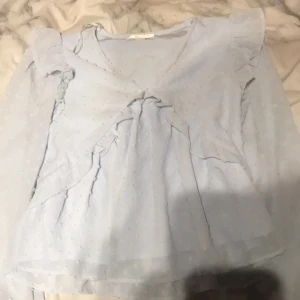 Ljusblå blus från Gina Tricot - Säljer en söt ljusblå blus från Gina Tricot med volangdetaljer och v-ringning. Blusen har långa ärmar och ett diskret prickigt mönster. Perfekt för en stilren look!