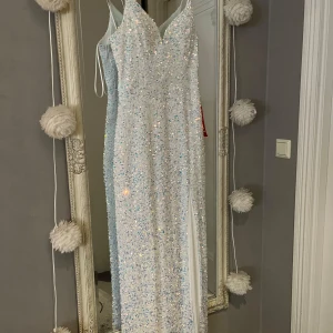 Vit glittrig lång balklänning - Säljer en fantastisk vit långklänning med glittrande paljetter och smala axelband. Klänningen har en elegant slits och en v-ringad design som ger en glamorös look. Perfekt för speciella tillfällen! Den är aldrig använd och nypris 3500