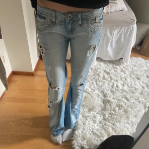 Ljusa slitna bootcut jeans - Säljer ett par ljusa bootcut jeans med slitningar på låren och knäna. De har en klassisk femficksdesign och är lågmidjade. Perfekta för en avslappnad stil med en vintage touch. Utsydda i benen