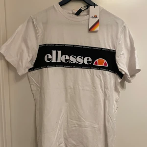 Vit t-shirt från Ellesse - Säljer en vit t-shirt från Ellesse med ett svart band och logotyp över bröstet. T-shirten är kortärmad och har en klassisk rund halsringning. Perfekt för en avslappnad stil. Oanvänd och etikett finns kvar.